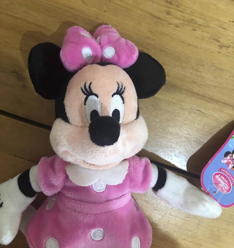 May include: Pink plush Minnie Mouse toy wearing a pink polka dot dress and a pink bow with white polka dots. The toy has black eyes, a black nose, and a white face. The toy is sitting on a wooden surface. The tag says "Disney Store Exclusive".