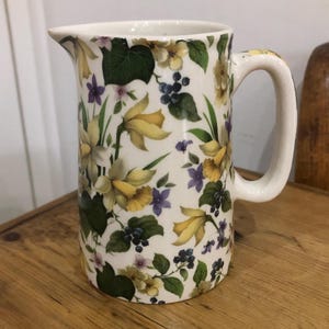 Può includere: Un'anfora in ceramica bianca con un motivo floreale con narcisi gialli, fiori viola, foglie verdi e bacche bianche.