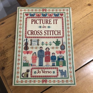 May include: A book titled "Picture It in Cross Stitch" by Jo Verso. The cover features a colourful cross-stitch design of a family, a cat, a dog, and various other objects.