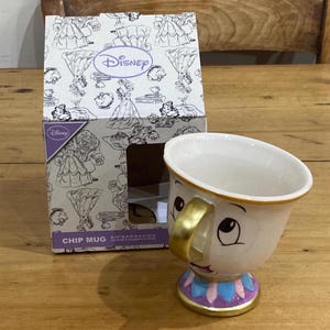 May include: A Chip mug from Disney's Beauty and the Beast, with a gold handle and base. The mug has a white body with Chip's face and a purple, blue, and gold base. The mug is next to a Disney-branded box.