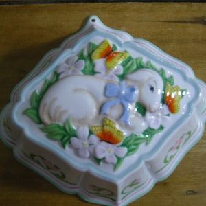 May include: A white ceramic mould shaped like a lamb with a blue ribbon around its neck. The mould is surrounded by green leaves and pink and yellow flowers.