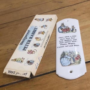Op de afbeelding: Een vintage Peter Rabbit set met een keramische wanddecoratie en een doos. Het witte keramische stuk toont illustraties en tekst uit het verhaal. De doos is crèmekleurig met illustraties van Peter Rabbit en de tekst "Wedgwood PETER RABBIT".