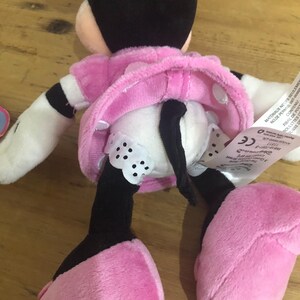 May include: A plush toy of Minnie Mouse wearing a pink dress with white polka dots and black shoes. The toy has a black tail and a black bow on her head. The toy is sitting on a wooden surface.