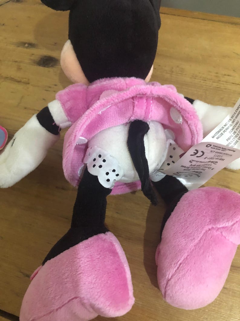 May include: A plush toy of Minnie Mouse wearing a pink dress with white polka dots and black shoes. The toy has a black tail and a black bow on her head. The toy is sitting on a wooden surface.