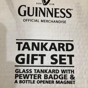 May include: A white box with black text that reads "GUINNESS OFFICIAL MERCHANDISE TANKARD GIFT SET GLASS TANKARD WITH PEWTER BADGE & A BOTTLE OPENER MAGNET". The Guinness logo is in the top left corner.