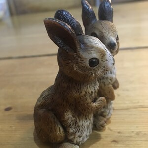 May include: Two brown and white ceramic rabbit figurines. The rabbits are standing upright with their ears perked up. The rabbit on the left is slightly larger than the rabbit on the right.