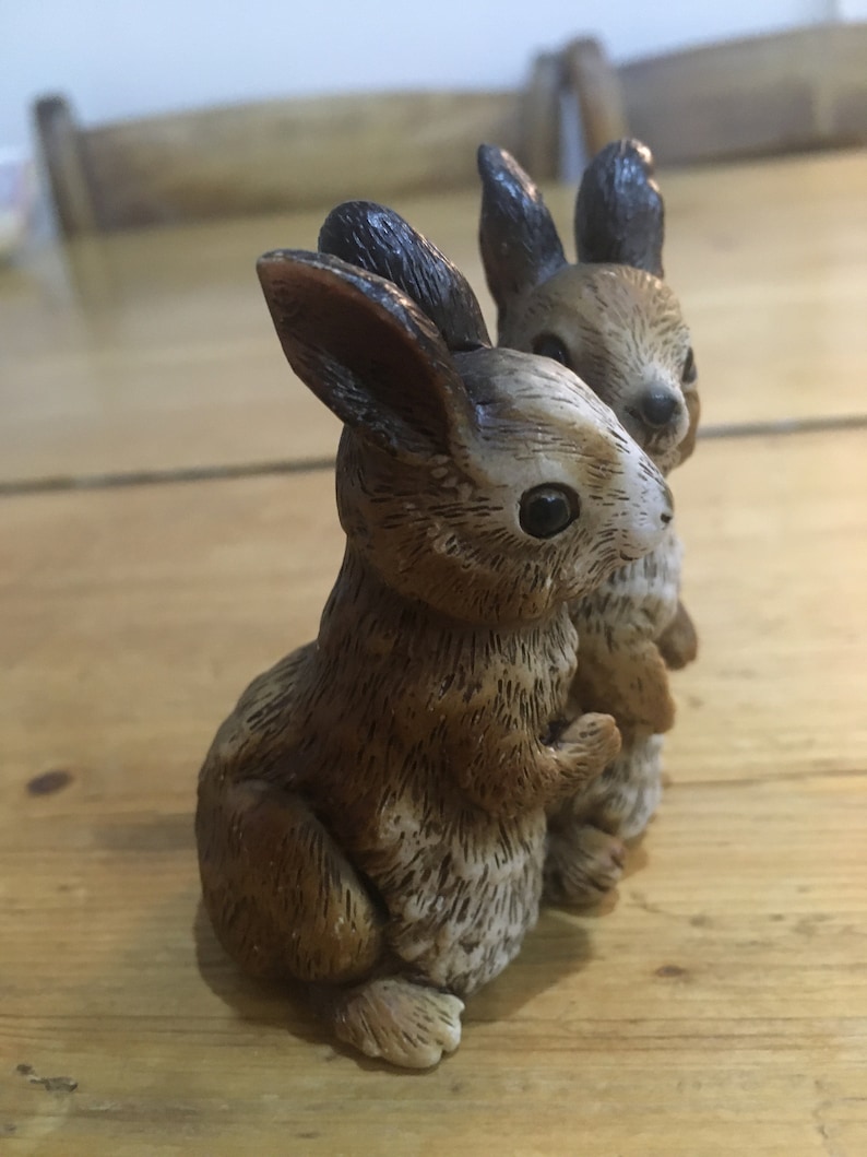 May include: Two brown and white ceramic rabbit figurines. The rabbits are standing upright with their ears perked up. The rabbit on the left is slightly larger than the rabbit on the right.