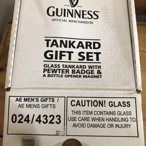 May include: A white box with black text that reads "Guinness Tankard Gift Set". The box contains a glass tankard, a pewter badge, and a bottle opener magnet. The box also has a caution label that reads "Caution! Glass. This item contains glass. Use care when handling to avoid damage or injury."