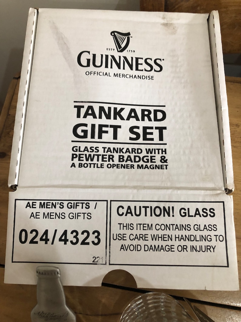 May include: A white box with black text that reads "Guinness Tankard Gift Set". The box contains a glass tankard, a pewter badge, and a bottle opener magnet. The box also has a caution label that reads "Caution! Glass. This item contains glass. Use care when handling to avoid damage or injury."