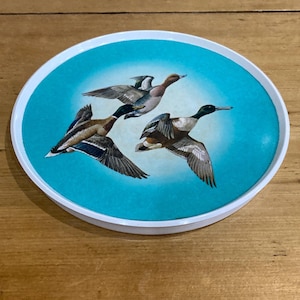 May include: Round coaster with a white rim and a teal center featuring three ducks in flight. The ducks have brown, black, and white feathers, with wings spread wide. The coaster is likely made of ceramic or a similar material.