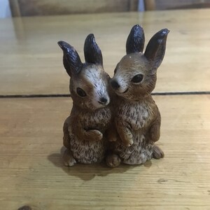 May include: Two brown ceramic rabbit figurines with black ears and brown fur. The rabbits are facing each other and appear to be cuddling.