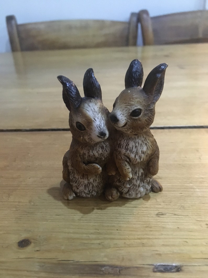 May include: Two brown ceramic rabbit figurines with black ears and brown fur. The rabbits are facing each other and appear to be cuddling.