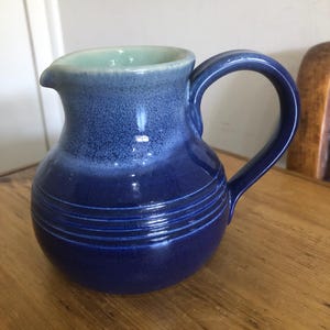 Op de afbeelding: Een kleine keramische kan met een afgeronde body en een gebogen handvat. De kan is geglazuurd in blauwtinten, met een lichtere blauwe binnenkant en een donkerdere blauwe buitenkant. Horizontale lijnen omcirkelen de basis.