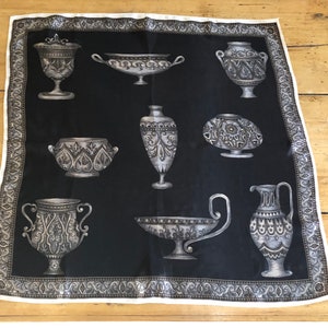 Puede incluir: Un pañuelo de seda negro con un borde blanco y un patrón repetido de jarrones y cuencos griegos plateados. Los jarrones y los cuencos están decorados con intrincados diseños florales.