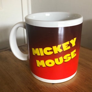 Puede incluir: Taza de cer&aacute;mica blanca con un fondo degradado rojo y marr&oacute;n. Las palabras "Mickey Mouse" est&aacute;n impresas en amarillo en la taza.