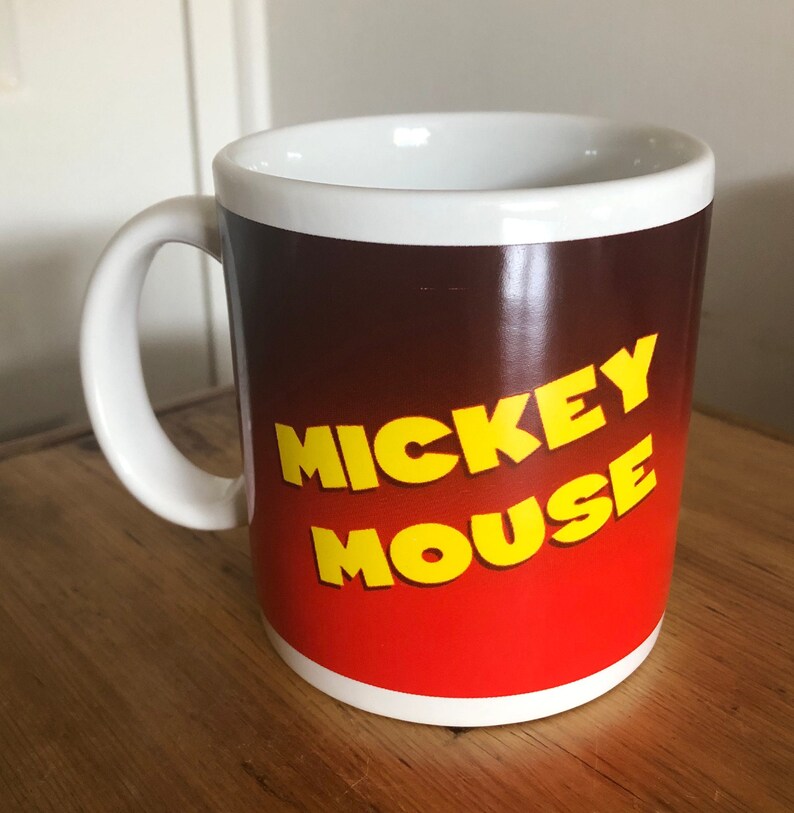 Puede incluir: Taza de cer&aacute;mica blanca con un fondo degradado rojo y marr&oacute;n. Las palabras "Mickey Mouse" est&aacute;n impresas en amarillo en la taza.