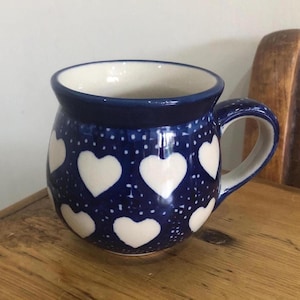 Pode incluir: Uma caneca de cerâmica com exterior azul escuro adornada com formas de coração brancas e pequenos pontos brancos. A caneca tem um corpo arredondado, interior branco e alça azul escuro. Está sobre uma superfície de madeira.
