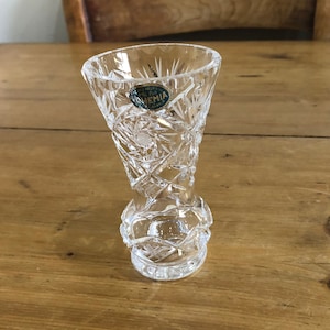 May include: A clear glass vase with a cut crystal design. The vase has a round base and a tall, slender neck. A green label with the word "Bohemia" is attached to the vase.
