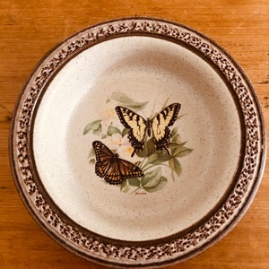 May include: Round ceramic plate with a speckled cream-colored center and a brown patterned border. The plate features a butterfly and floral design in the center, with a yellow and black butterfly and a brown and black butterfly.