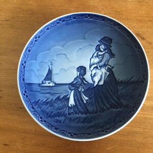 Op de afbeelding: Blauw en wit porseleinen bord met een scène van drie vrouwen en een zeilboot in de verte. Het bord heeft een blauwe en witte rand.