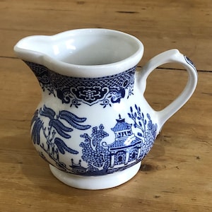 Puede incluir: Una jarra de crema de cerámica blanca con un patrón de sauce azul y blanco. La jarra tiene un asa y un pico.