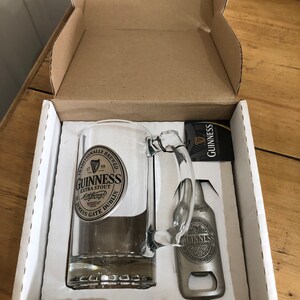 May include: A white box with a Guinness beer glass and bottle opener. The glass has a silver Guinness logo and the bottle opener has a silver Guinness logo.