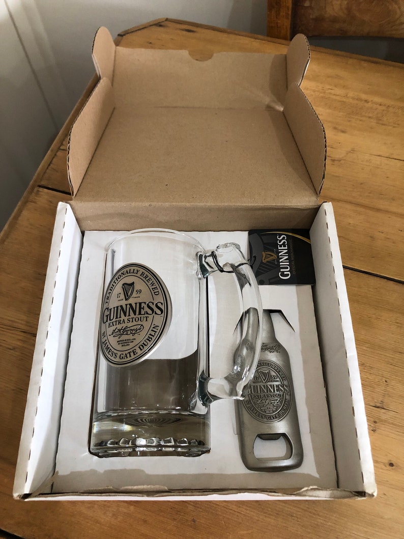 May include: A white box with a Guinness beer glass and bottle opener. The glass has a silver Guinness logo and the bottle opener has a silver Guinness logo.