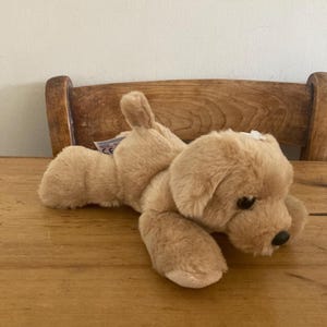 Puede incluir: Un perro de peluche color canela con pelaje suave, orejas caídas y una pequeña cola erguida. El juguete tiene ojos de plástico marrón oscuro y una nariz negra, con una pequeña etiqueta blanca cerca de la cola y otra etiqueta blanca en la cabeza.