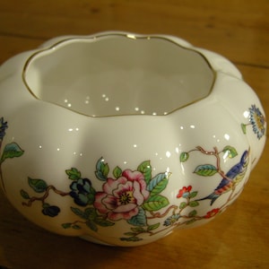 May include: A white ceramic bowl with a scalloped rim and a floral design. The bowl features a pink rose, blue flowers, green leaves, and a blue bird.