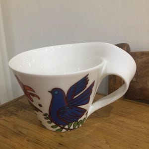 Puede incluir: Taza de té de cerámica blanca con un diseño de asa único. La taza presenta una ilustración de un pájaro azul con detalles en rojo y verde. La taza está sobre una superficie de madera.