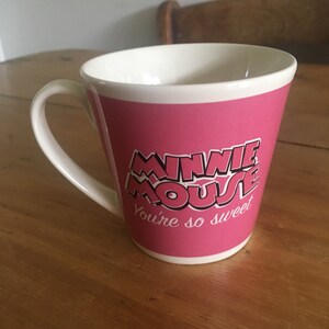 Peut inclure: Une tasse en c&eacute;ramique rose avec un int&eacute;rieur et une anse blancs. La tasse porte les mots "MINNIE MOUSE" dans une police stylis&eacute;e, avec "You're so sweet" en dessous. La tasse est pos&eacute;e sur une surface en bois.