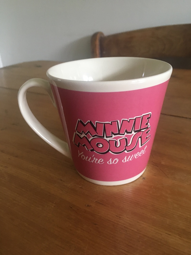 Peut inclure: Une tasse en c&eacute;ramique rose avec un int&eacute;rieur et une anse blancs. La tasse porte les mots "MINNIE MOUSE" dans une police stylis&eacute;e, avec "You're so sweet" en dessous. La tasse est pos&eacute;e sur une surface en bois.