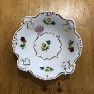 Puede incluir: Un tazón de porcelana blanca con un borde dorado y diseños florales. El tazón tiene un borde festoneado y una pequeña pegatina rosa con la palabra "Lux" en ella.