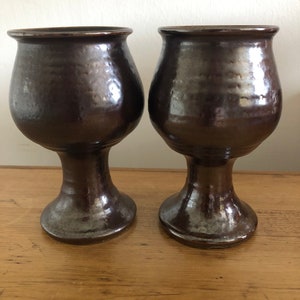 Puede incluir: Dos copas de cerámica marrón oscuro con un brillo metálico. Las copas tienen una copa ancha y un tallo alto y delgado.