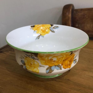 Könnte beinhalten: Eine weiße Keramikschüssel mit grünem Rand und einem floralen Muster aus gelben Rosen und grünen Blättern.