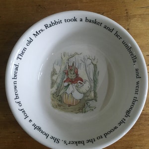 Könnte beinhalten: Eine weiße Keramikschüssel mit einem Bild eines Kaninchens, das einen Korb in einem Wald trägt. Der Rand der Schüssel ist mit dem Text "She bought a loaf of brown bread. Then old Mrs. Rabbit took a basket and her umbrella, and went through the wood to the baker's." beschriftet.