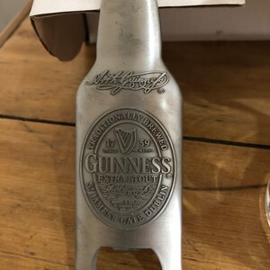 May include: Silver bottle opener shaped like a Guinness beer bottle. The bottle opener has the Guinness logo and text: "Traditionally Brewed", "17 Trade Mark", "59 TM Mark", "Guinness", "Extra Stout", "Arthur Guinness", "St. James's Gate Dublin".