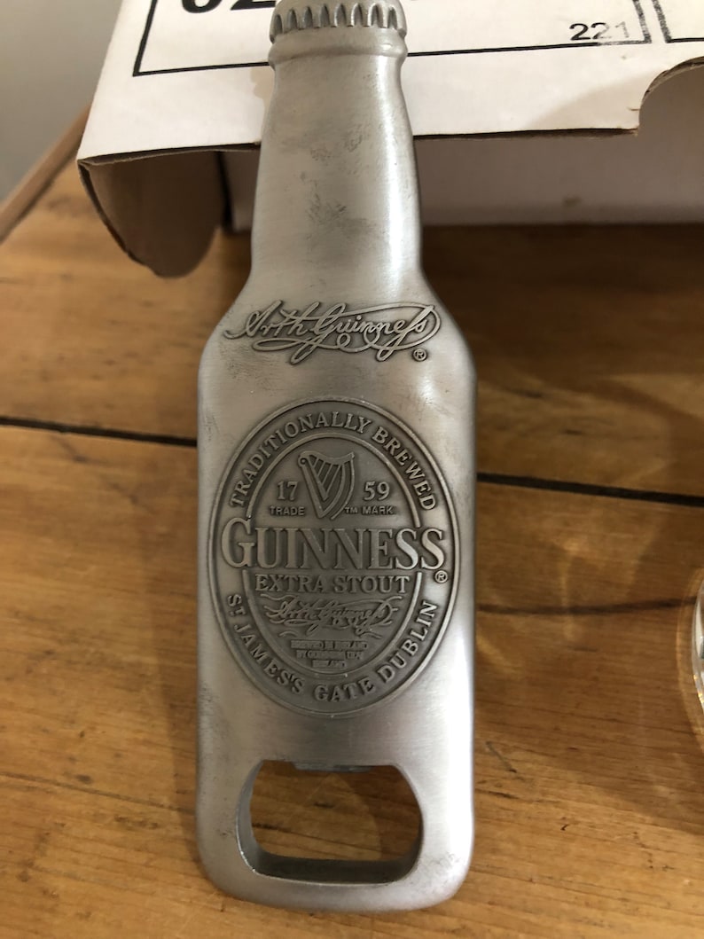 May include: Silver bottle opener shaped like a Guinness beer bottle. The bottle opener has the Guinness logo and text: "Traditionally Brewed", "17 Trade Mark", "59 TM Mark", "Guinness", "Extra Stout", "Arthur Guinness", "St. James's Gate Dublin".