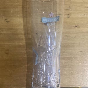 Op de afbeelding: Een helder, hoog bierglas met het Heineken-logo en sterontwerp. Het glas heeft een licht uitlopende vorm en is gemaakt van transparant glas. Het Heineken-logo is in blauw en roze. Het glas is leeg.