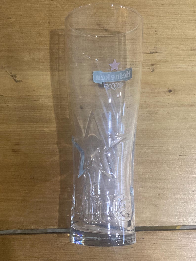 Op de afbeelding: Een helder, hoog bierglas met het Heineken-logo en sterontwerp. Het glas heeft een licht uitlopende vorm en is gemaakt van transparant glas. Het Heineken-logo is in blauw en roze. Het glas is leeg.