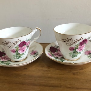 Può includere: Due tazze da tè bianche e piattini con un motivo floreale rosa e rosso. Le tazze hanno un bordo dorato e riportano la scritta "Ruby Wedding".