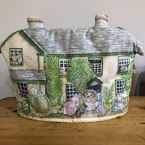 Può includere: Un copriteiera di colore crema a forma di casa con bordi verdi e tetto grigio. La casa ha finestre con piccole figure che guardano fuori. Il copriteiera è decorato con una scena da giardino con fiori e animali.