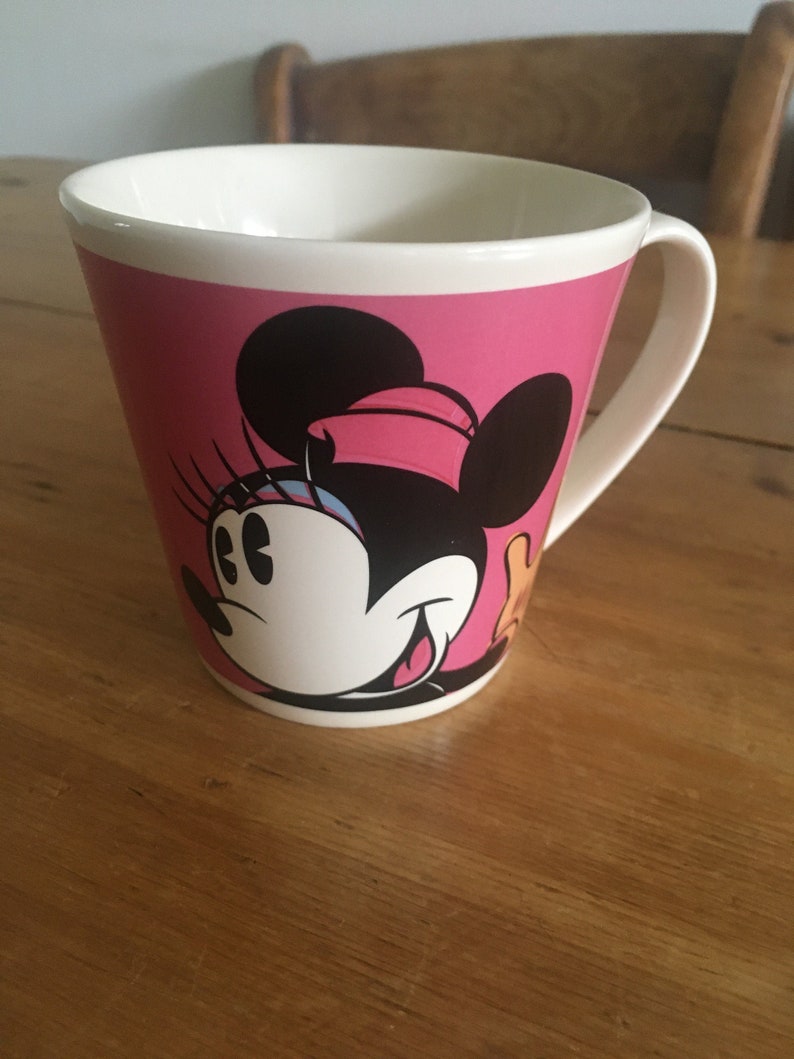 Peut inclure: Tasse en c&eacute;ramique rose avec un dessin de Minnie Mouse. L'int&eacute;rieur et la anse sont blancs. Le dessin de Minnie Mouse comprend des oreilles noires, un n&oelig;ud rose et un visage blanc. La tasse est pos&eacute;e sur une table en bois.