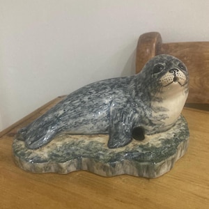 Puede incluir: Escultura de cerámica de una foca, tumbada sobre una base texturizada. La foca tiene un cuerpo gris y blanco con una cara detallada. La base es de color verde y gris moteado, dando la impresión de un hábitat natural. Es un objeto decorativo.
