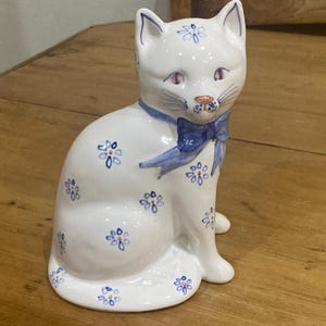 May include: A white ceramic cat figurine with blue floral accents and a blue bow. The cat is sitting upright with its ears perked up. The figurine is decorated with small blue flower patterns.