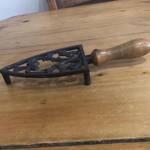 May include: A black cast iron trivet with a floral design and a wooden handle. The trivet has three small legs. The handle is light brown and oval-shaped. This antique item is likely used for placing hot items, such as a hot iron.