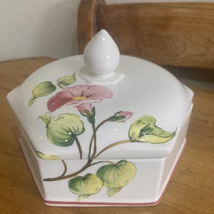 May include: A white ceramic hexagonal trinket box with a lid, featuring a floral design of pink flowers and green leaves. The box has a small white knob on top and a red trim around the base.