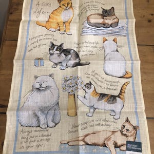 以下が含まれることがあります： ベージュ色のアイリッシュリネンティータオル。さまざまなポーズの猫のイラストが描かれています。タオルには猫に関連するフレーズや引用文が書かれており、薄い青色の縁取りがあります。