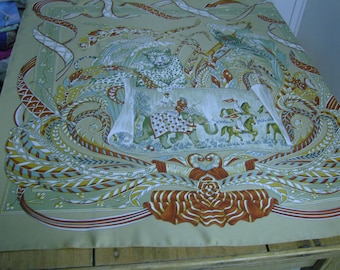 May include: A beige silk scarf with a colourful design featuring a leopard, an elephant, and riders on horseback. The design is surrounded by intricate patterns of ribbons and leaves.