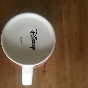 Peut inclure: Mug en c&eacute;ramique blanche avec une anse, portant le logo Disney et le symbole du copyright. Le mug est pos&eacute; sur une surface en bois. L'int&eacute;rieur du mug est blanc.